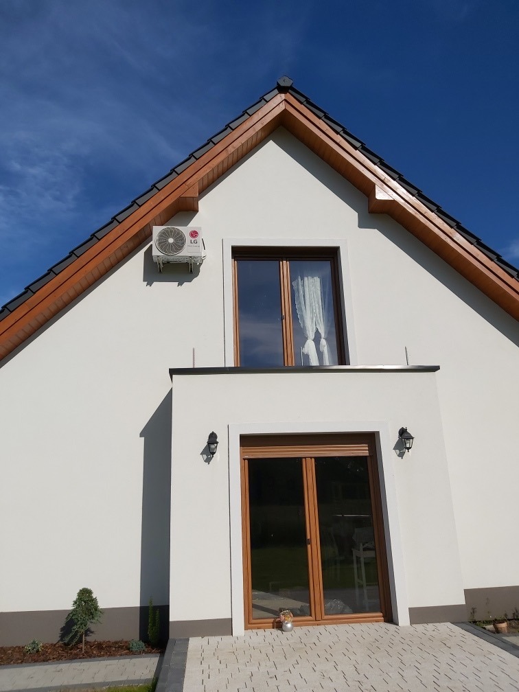 Biały dom z zamontowaną klimatyzacją LG na ścianie, Ząbkowice Śląskie. Widoczny fragment dachu z brązowymi elementami i okno z białą firanką.