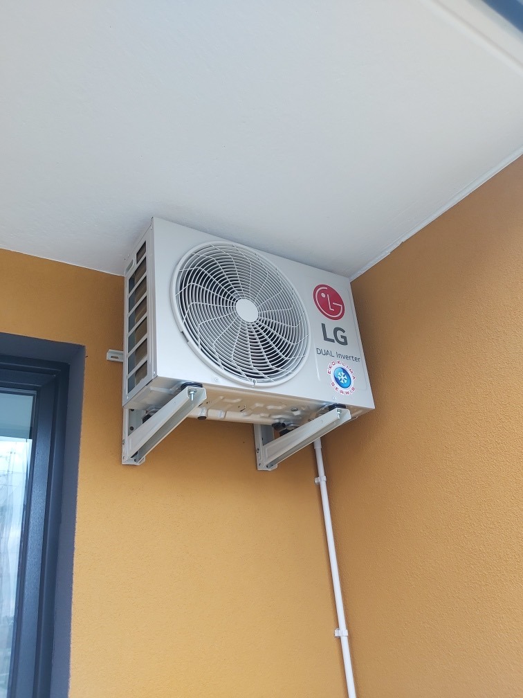 Biała jednostka zewnętrzna klimatyzatora LG Dual Inverter zamontowana na żółtej ścianie budynku, widoczne białe rury instalacyjne.