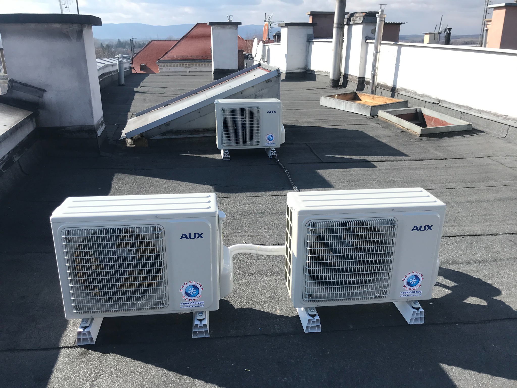 Trzy jednostki zewnętrzne klimatyzacji marki AUX zamontowane na płaskim dachu budynku z widocznymi kominami i panoramą miasta w tle.