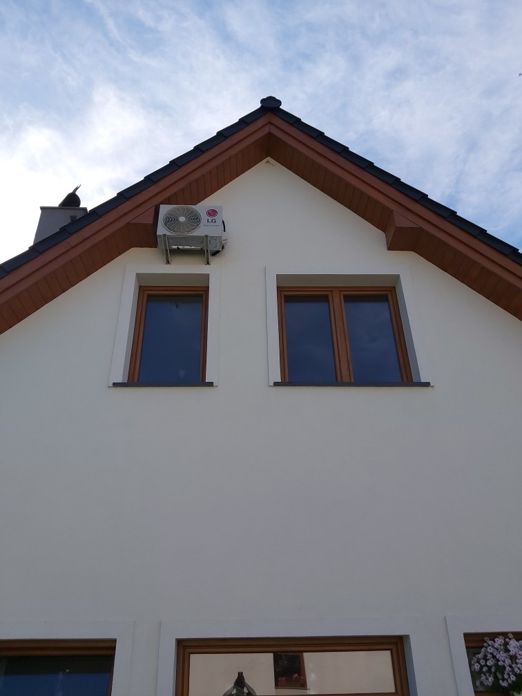 Biała fasada domu z brązowymi oknami i zamontowaną jednostką zewnętrzną klimatyzatora LG pod okapem dachu, widok z dołu.