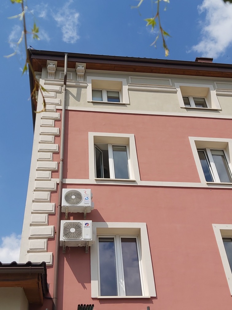 Dwa klimatyzatory zamontowane na elewacji budynku w kolorze pudrowego różu, widoczne okna z białymi ramami i ozdobne elementy architektoniczne, błękitne niebo w tle.