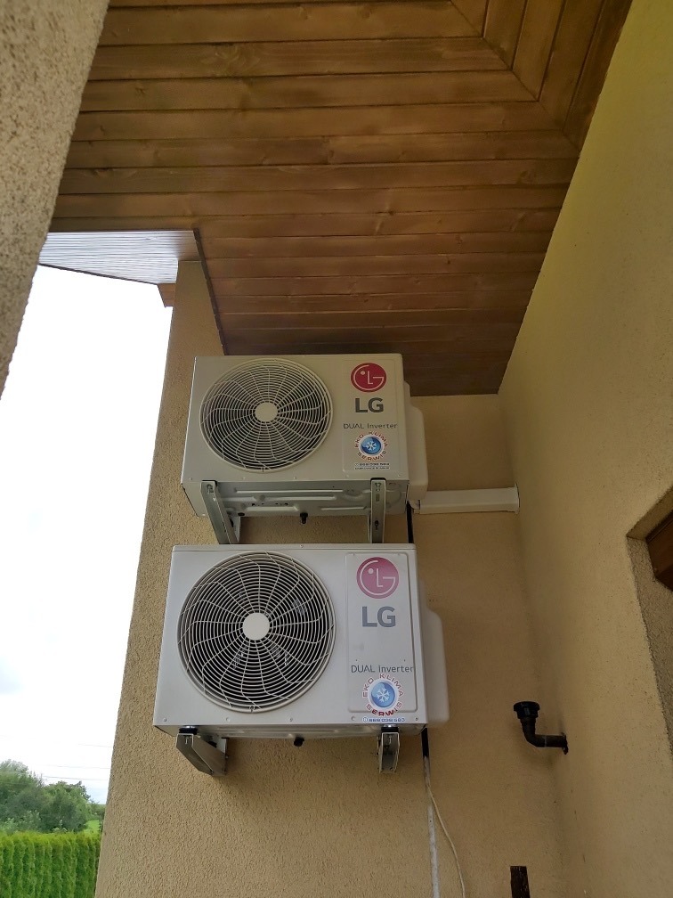 Dwa klimatyzatory LG Dual Inverter zamontowane jeden nad drugim na beżowej elewacji budynku pod drewnianym okapem.