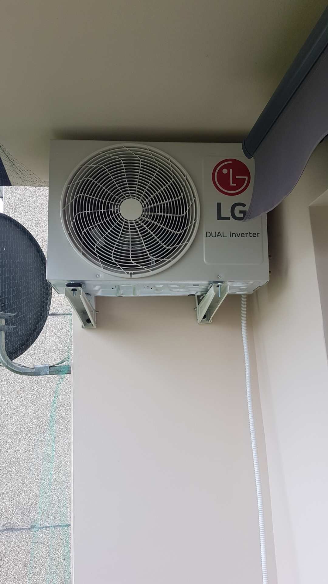 Biała jednostka zewnętrzna klimatyzatora LG Dual Inverter zamontowana na ścianie budynku, widoczne wsporniki montażowe i peszel z kablem.