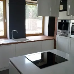 Jasna kuchnia z białymi szafkami, drewnianym blatem pod oknem i wyspą kuchenną z płytą indukcyjną, widok na zewnątrz przez okno z brązową ramą.