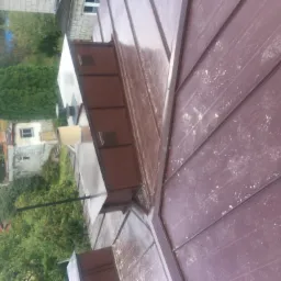 Nowy, brązowy dach z blachy trapezowej, z widocznymi obróbkami blacharskimi komina i śladami zabrudzeń, na tle zieleni i zabudowań.