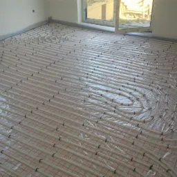 Białe rurki ogrzewania podłogowego, ułożone w pętle na folii z czerwoną siatką, widok z góry, okno w tle.