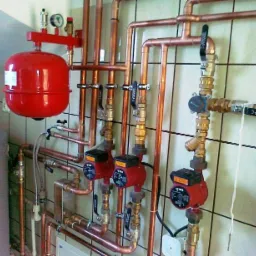 Złożona instalacja centralnego ogrzewania z miedzianych rur, czerwonym zbiornikiem wyrównawczym i trzema pompami obiegowymi na tle kafelkowanej ściany.