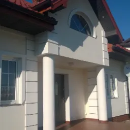 Elewacja domu jednorodzinnego z kolumnami i czerwonym dachem na tle błękitnego nieba. Widoczne detale architektoniczne i schody wejściowe.