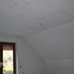 Poddasze w trakcie wykańczania: surowy sufit i ściany po nałożeniu gładzi, widoczne ślady szpachlowania, wystające przewody oświetleniowe, okno z widokiem na zieleń.