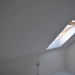 Poddasze z oknem dachowym, ściany i sufit pokryte wstępnie przygotowaną powierzchnią pod gładzie, widoczne niedoskonałości na suficie. Przygotowane podłoże pod wykończenie.