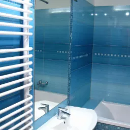 Niebieska łazienka z białymi akcentami: umywalka z baterią, grzejnik drabinkowy, lustro z oświetleniem i wanna w tle, wykończona mozaiką.