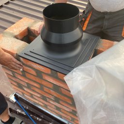 Zakład instalacyjny Pazoła Janusz - Montaż czarnego komina na ceglanym kominie na dachu z blachy trapezowej. Widoczne dłonie pracownika w ubraniu roboczym z pomarańczowymi wstawkami. Częściowo zakryty folią ochronną.