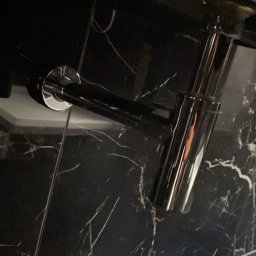 Zakład instalacyjny Pazoła Janusz - Sifon umywalkowy z chromowanego metalu na tle czarnej marmurowej ściany z białymi żyłkami.