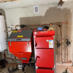 Zakład instalacyjny Pazoła Janusz - Czerwony piec na paliwo stałe Defro z zasobnikiem, podłączony do instalacji grzewczej z miedzianymi rurami i białym zbiornikiem na wodę, widoczny fragment podłogi i ściany w surowym wykończeniu.