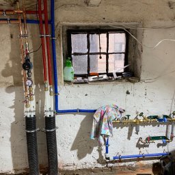 Zakład instalacyjny Pazoła Janusz - Instalacja wodna z rur miedzianych i plastikowych w piwnicy, widoczne zawory, manometr, rury w peszlach, okno z narzędziami i butelką, ściana z niedoskonałościami.