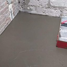 Zakład instalacyjny Pazoła Janusz - Wylewka betonowa w pomieszczeniu z surowymi, częściowo otynkowanymi ścianami z cegły i betonu, widoczne narzędzia do rozprowadzania betonu.