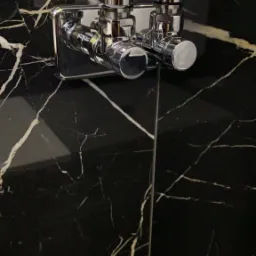 Błyszcząca, chromowana bateria umywalkowa z dwoma kurkami zamontowana na ścianie z czarnego marmuru z białymi żyłami.