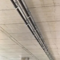 Rzędy rur instalacyjnych, prawdopodobnie wentylacyjnych, przymocowanych metalowymi uchwytami do surowego, betonowego stropu w surowym wnętrzu budynku w trakcie budowy.