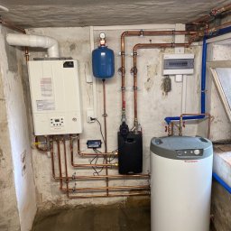 Zakład instalacyjny Pazoła Janusz - Nowoczesna instalacja grzewcza w piwnicy: biały piec gazowy Immergas, zasobnik ciepłej wody, niebieski zbiornik wyrównawczy i miedziane rury, całość zamontowana na surowych, betonowych ścianach.