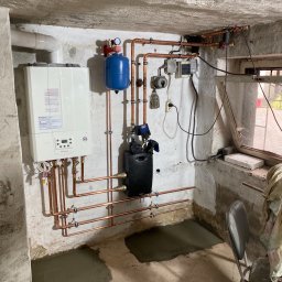 Zakład instalacyjny Pazoła Janusz - Nowy, biały piec gazowy Immergas zamontowany na ścianie w piwnicy, z widocznymi miedzianymi rurami, niebieskim zbiornikiem wyrównawczym, czarną pompą obiegową i świeżym betonem na podłodze.