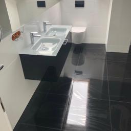 Zakład instalacyjny Pazoła Janusz - Nowoczesna łazienka z podwójną umywalką na czarnej szafce, białe płytki na ścianach, czarna armatura i lśniąca czarna podłoga z płytek, widoczne okablowanie w ścianie.