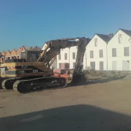 Żółta koparka CAT 318B na placu budowy z rzędami domów w tle, część w stanie surowym, część z gotową elewacją.