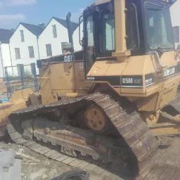 Żółty spychacz Caterpillar D5M LGP na placu budowy z nowymi domami w tle, widoczne elementy konstrukcyjne i opony.