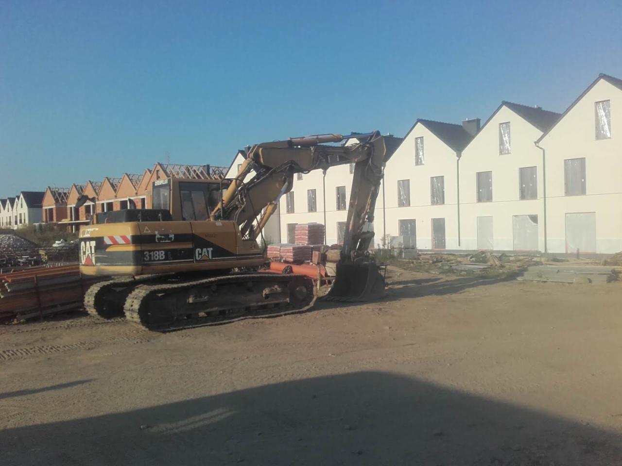 Żółta koparka CAT 318B na placu budowy z rzędami domów w tle, część w stanie surowym, część z gotową elewacją.