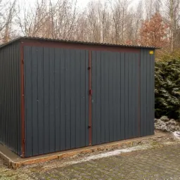 Gotowy, ciemnoszary garaż blaszany z dwuskrzydłowymi drzwiami, na betonowej wylewce, widok z boku na tle drzew i krzewów w pochmurny dzień.