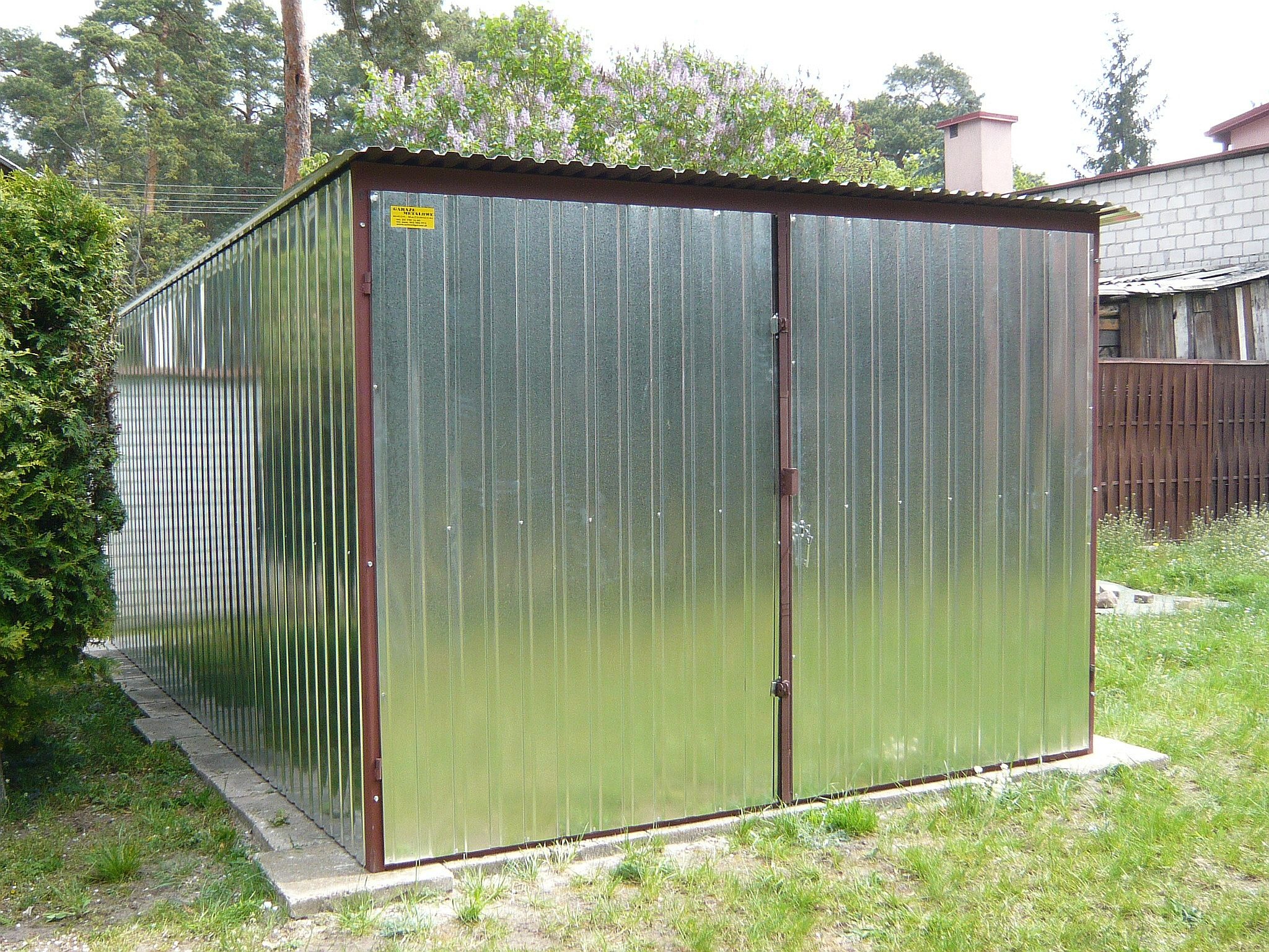 Gotowy garaż blaszany o wymiarach 3x5 metrów, z dwuskrzydłowymi drzwiami, ustawiony na betonowej podmurówce w ogrodzie.