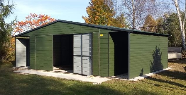 Zielony garaż blaszany z przesuwnymi drzwiami i otwartym bokiem, na betonowej wylewce, jesienne drzewa w tle.