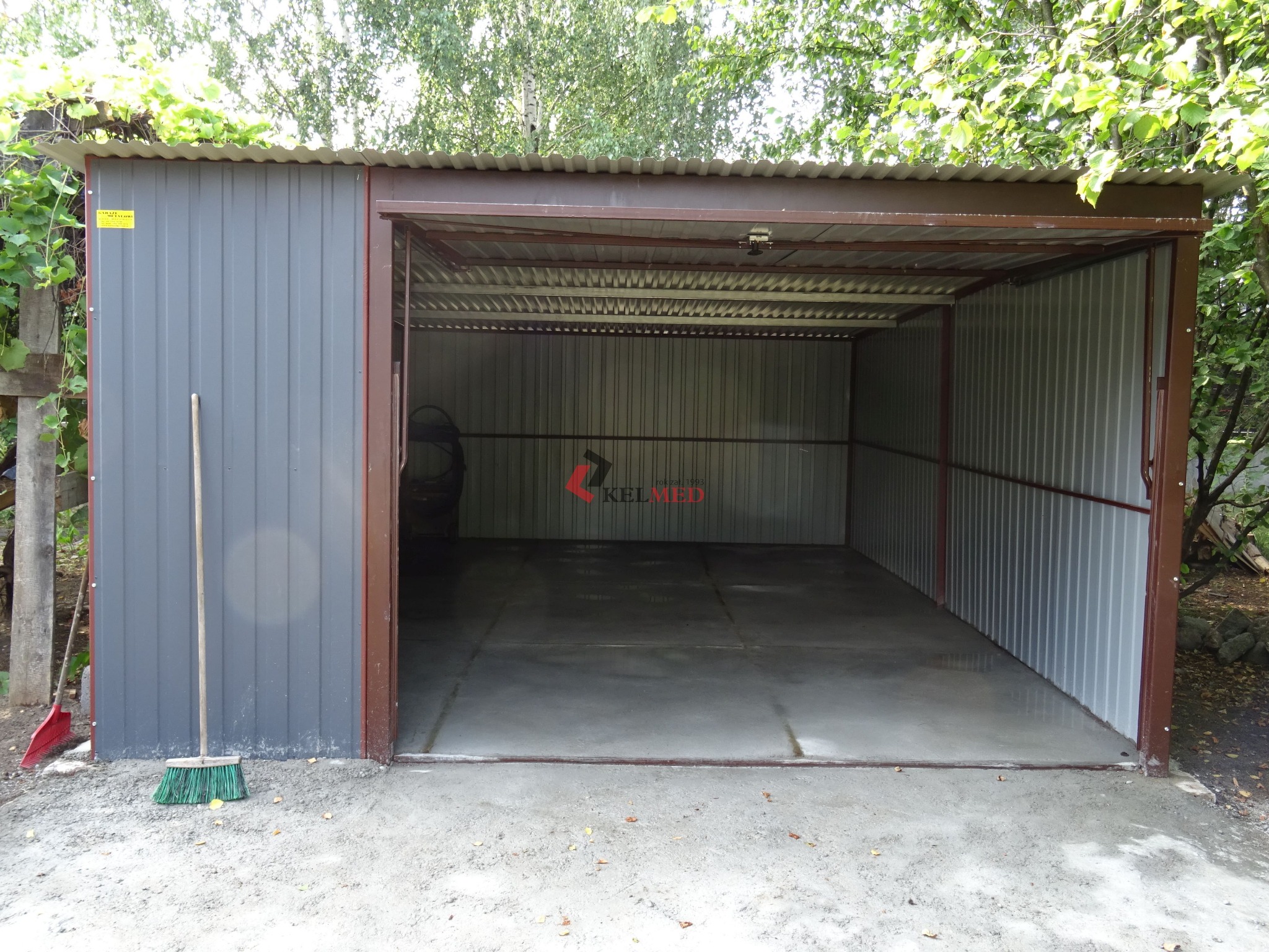 Otwarty, szary garaż z brązowymi elementami konstrukcyjnymi, widoczne wnętrze z betonową posadzką, na zewnątrz leży miotła.