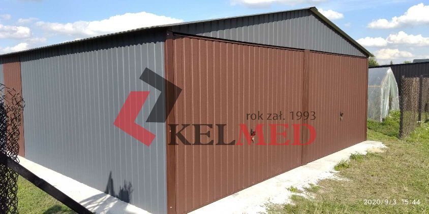 Gotowy garaż z blachy trapezowej w kolorze grafitowo-brązowym, z widocznym logo producenta na ścianie i datą założenia firmy, otoczony zielenią i szklarnią w tle.