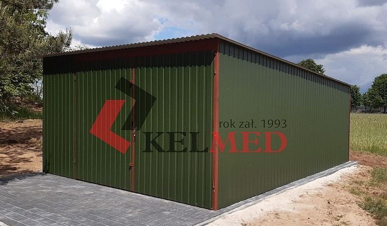 Zielony garaż blaszany z czerwonymi detalami, logo firmy 'KELMED' i rokiem założenia 1993, ustawiony na szarej kostce brukowej, na tle pola i drzew.