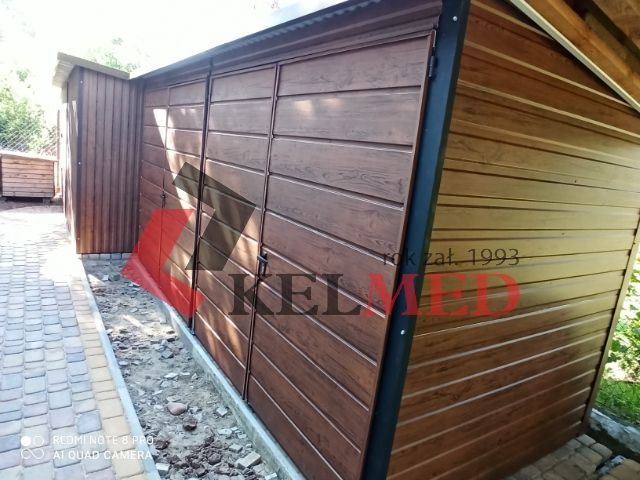 Brązowy garaż blaszany z poziomymi panelami, widok z boku, z widocznym fragmentem dachu i sąsiedniej drewnianej konstrukcji, teren wokół garażu jeszcze niezagospodarowany.