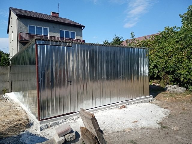 Nowo postawiony, metalowy garaż z blachy falistej, widoczny na betonowej wylewce przed domem jednorodzinnym w słoneczny dzień.