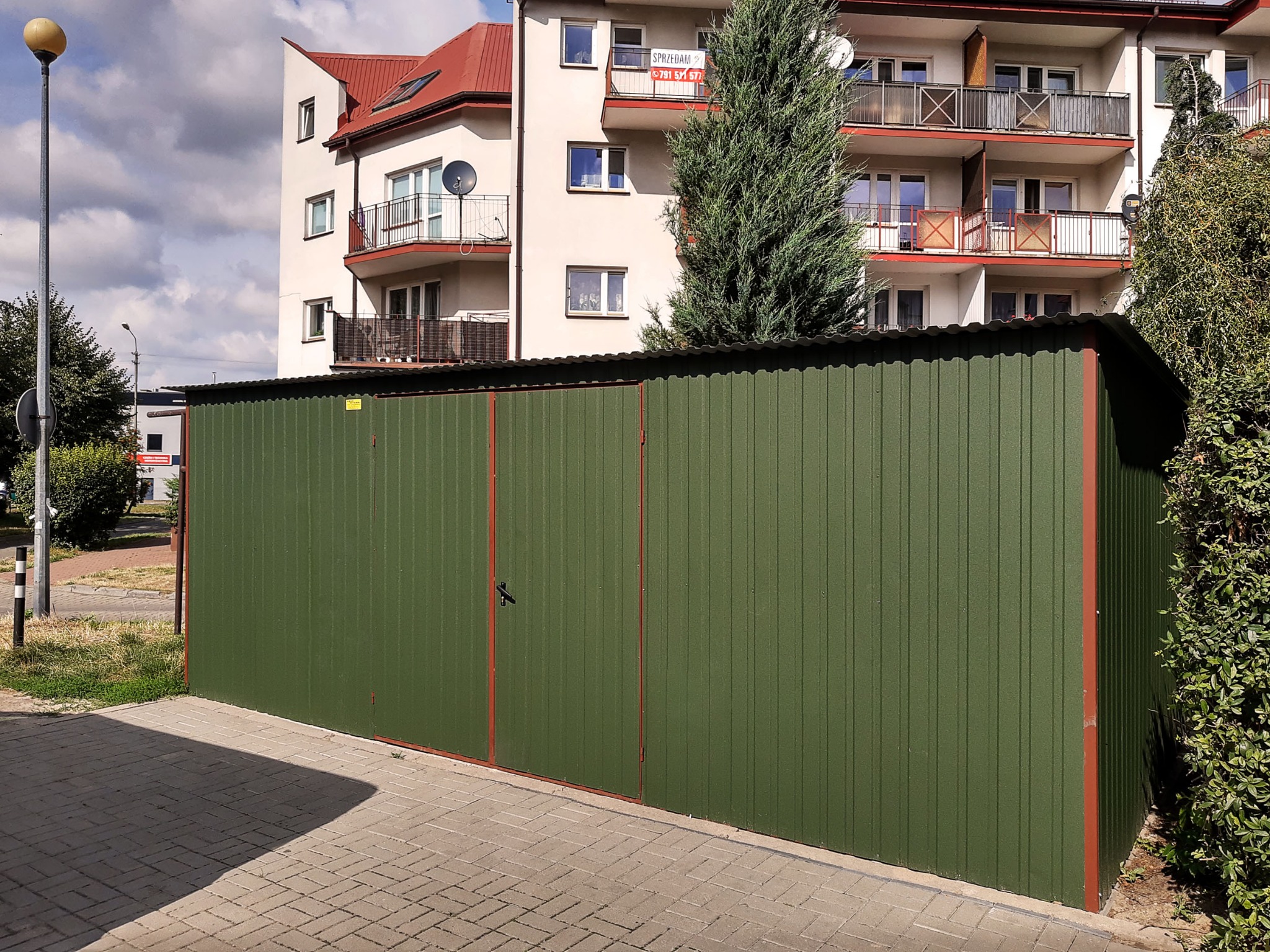 Zielony garaż blaszany z czerwoną ramą na tle bloku mieszkalnego z balkonami i drzewem iglastym.