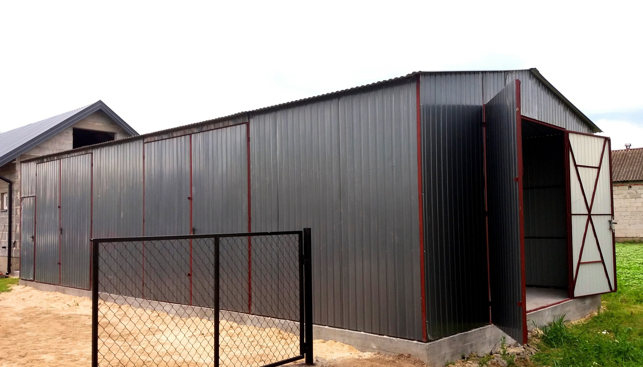 Szary, blaszany garaż z czerwoną ramą w Mińsku Mazowieckim, widoczny zza metalowego ogrodzenia. Jedne z wrót garażu są otwarte, ukazując białą konstrukcję wewnętrzną.