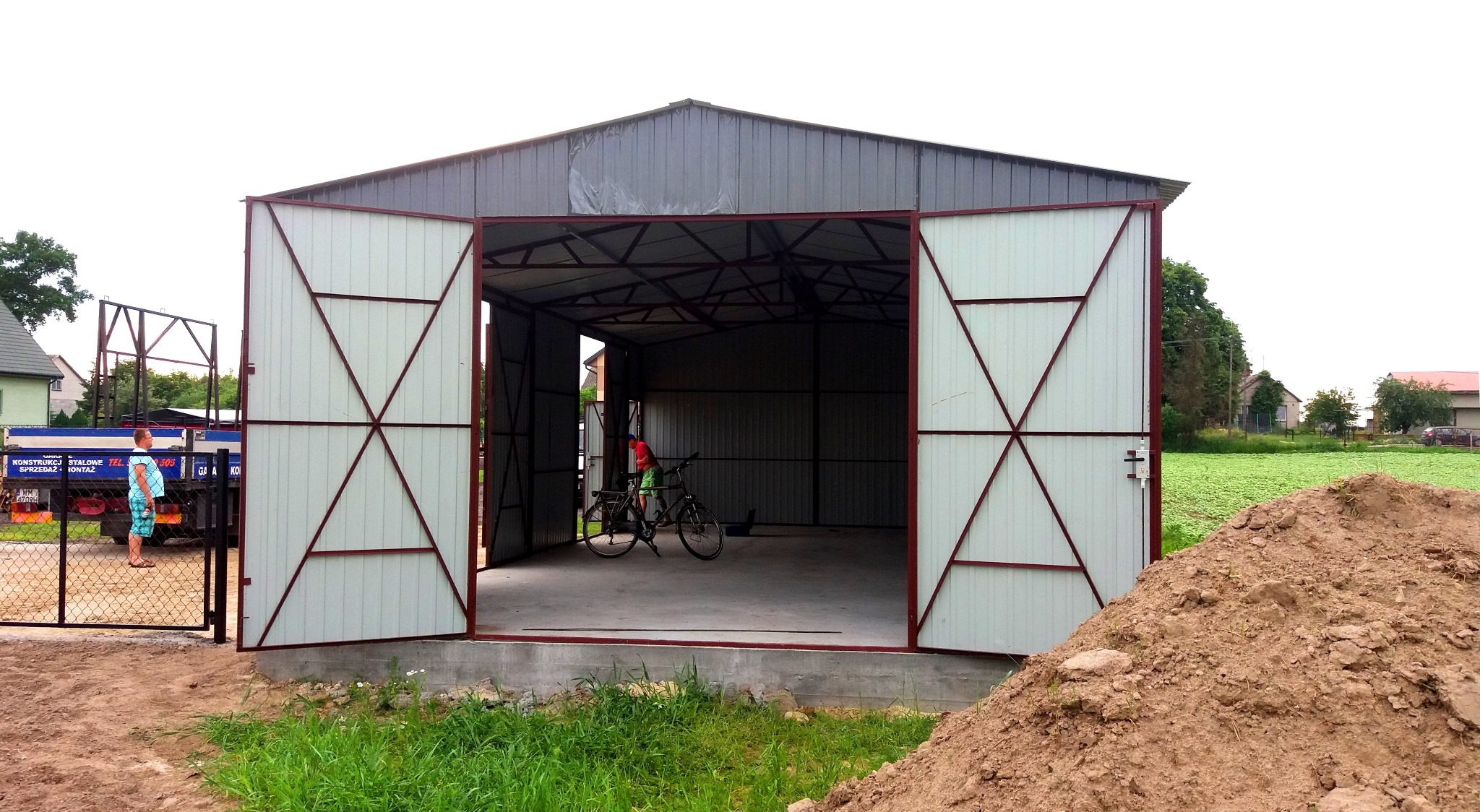 Otwarty, szary garaż z blachy trapezowej z białymi drzwiami z czerwonymi wzmocnieniami, wewnątrz rowery, na zewnątrz pryzma ziemi i częściowo widoczna ciężarówka z napisem 'konstrukcje stalowe'