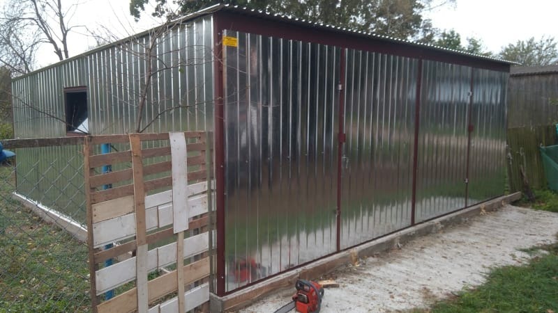 Gotowy garaż blaszany z oknem i dwuskrzydłowymi drzwiami, widoczny z boku, z paletą i piłą łańcuchową na pierwszym planie.