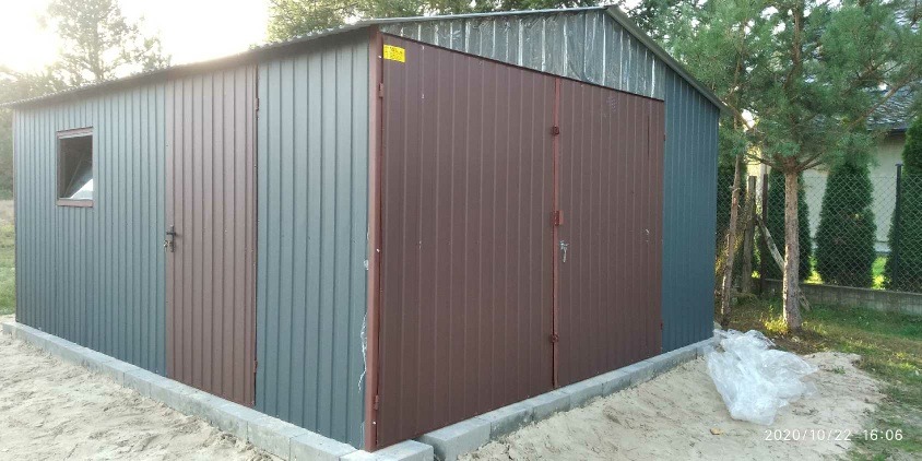 Zewnętrzny widok szarego garażu z blachy falistej z brązowymi drzwiami, osadzonego na betonowej podmurówce, z małym oknem po lewej stronie i drzewami w tle.