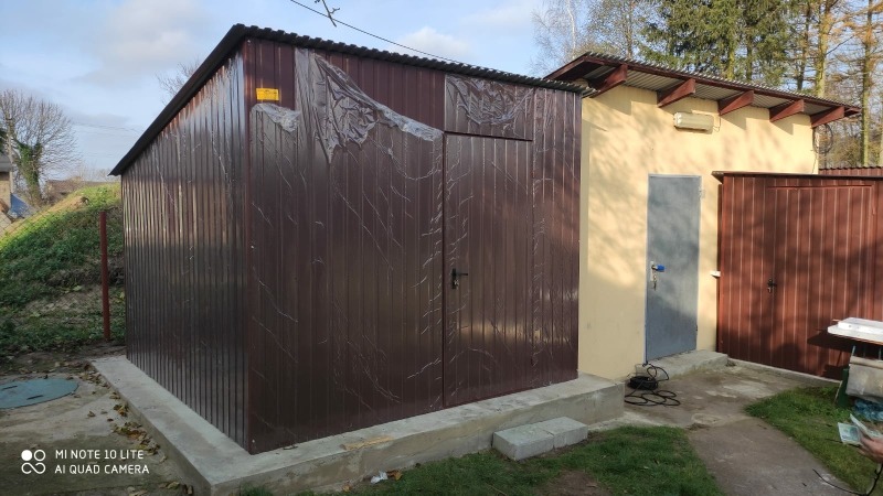 Brązowy garaż blaszany z dwuspadowym dachem, zabezpieczony folią ochronną na części powierzchni, stojący na betonowej wylewce obok budynku z szarą metalową furtką i brązową bramą.