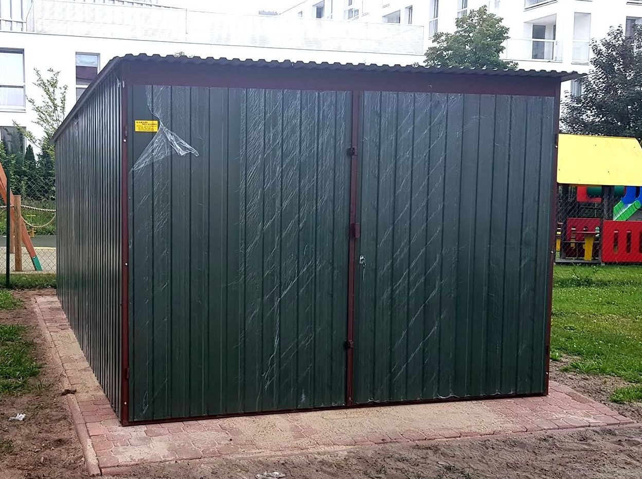 Zielony, dwudrzwiowy garaż z blachy falistej, ustawiony na chodniku z czerwonej kostki, z pozostałościami folii ochronnej na bocznej ścianie, na tle placu zabaw i bloku mieszkalnego.
