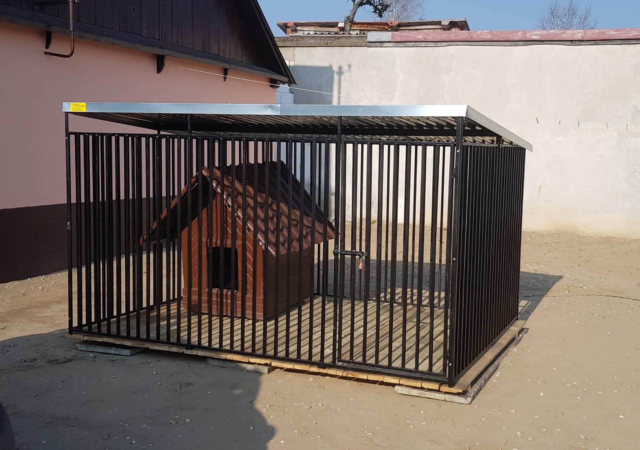 Klatka dla psa z zadaszeniem i budą w środku, umieszczona na drewnianych podkładach, na zewnątrz, w tle budynki.