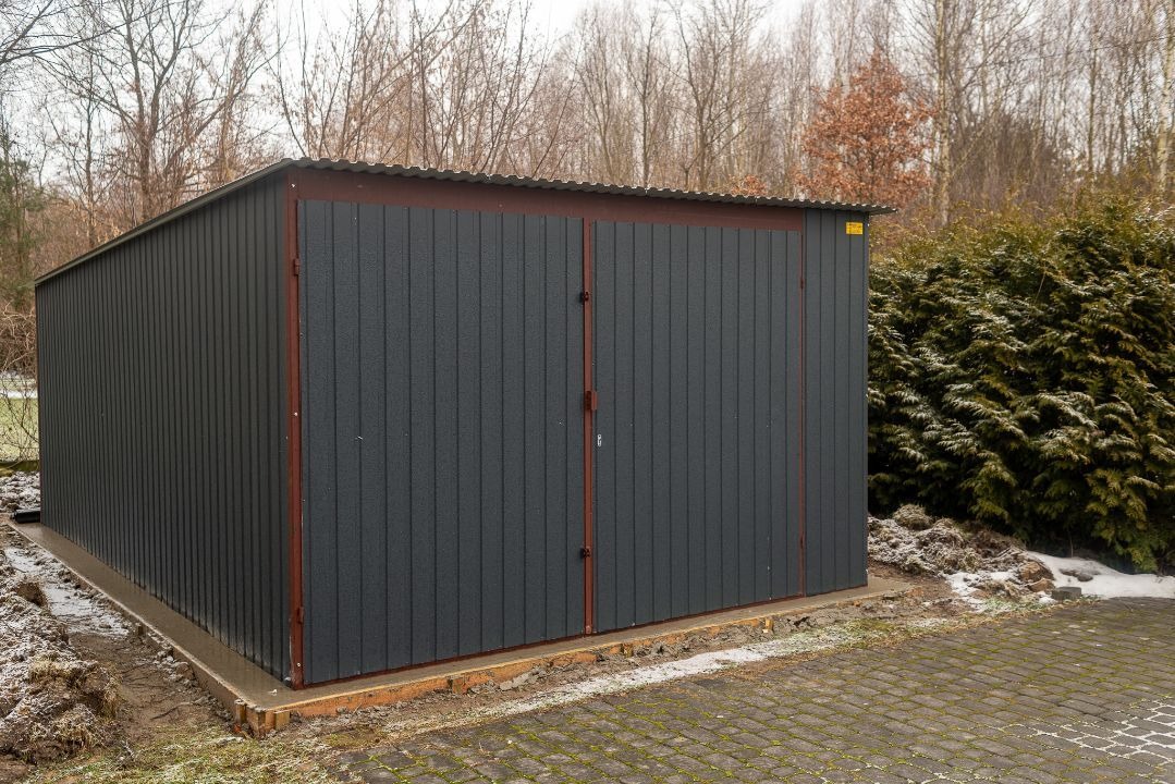 Gotowy, ciemnoszary garaż blaszany z dwuskrzydłowymi drzwiami, na betonowej wylewce, widok z boku na tle drzew i krzewów w pochmurny dzień.