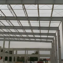 Szare, stalowe elementy konstrukcji hali w trakcie budowy, z widocznymi betonowymi słupami i fragmentami ścian. W tle zieleń i zabudowania.