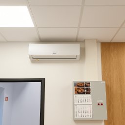 P.W. KLIMA Plus sp.j. - Biała klimatyzacja na ścianie w biurze, widoczny kalendarz i zegar. Kątowy kadr z otwartymi drzwiami po lewej stronie. Oświetlenie sufitowe.