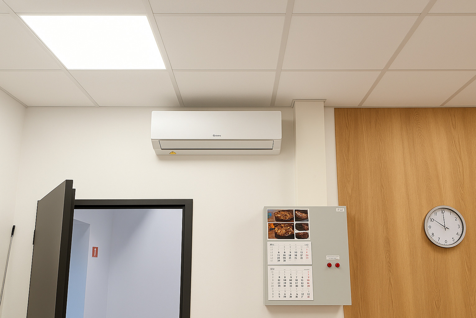 Biała klimatyzacja na ścianie w biurze, widoczny kalendarz i zegar. Kątowy kadr z otwartymi drzwiami po lewej stronie. Oświetlenie sufitowe.