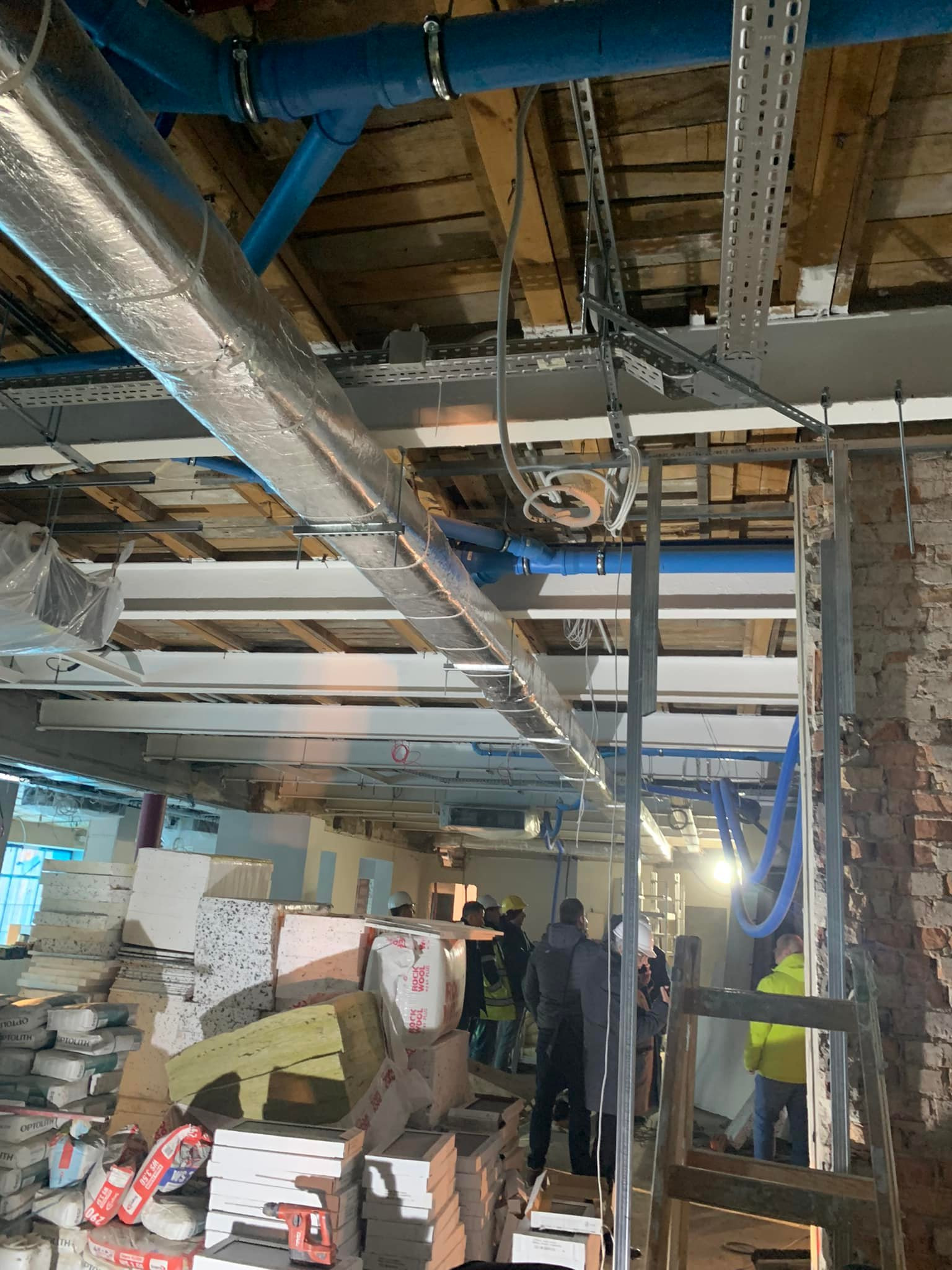 Montaż instalacji wentylacyjnej: rury wentylacyjne z izolacją, niebieskie rury, kable i drabina w surowym wnętrzu budynku w trakcie prac instalacyjnych.