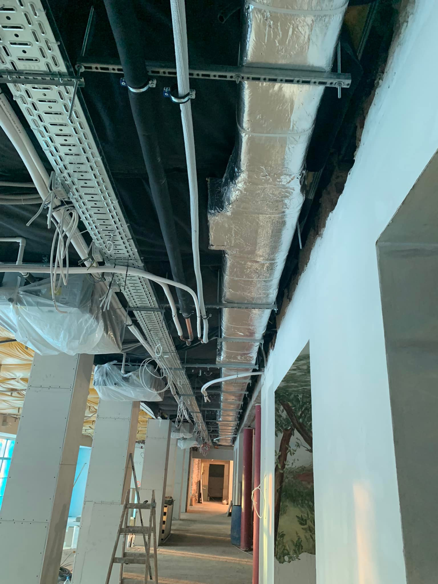 Instalacja wentylacji z folią aluminiową w budynku. Widoczne rury, kable, drabina i kolumny w trakcie wykończenia. Ujęcie z dołu, perspektywa zbieżna.